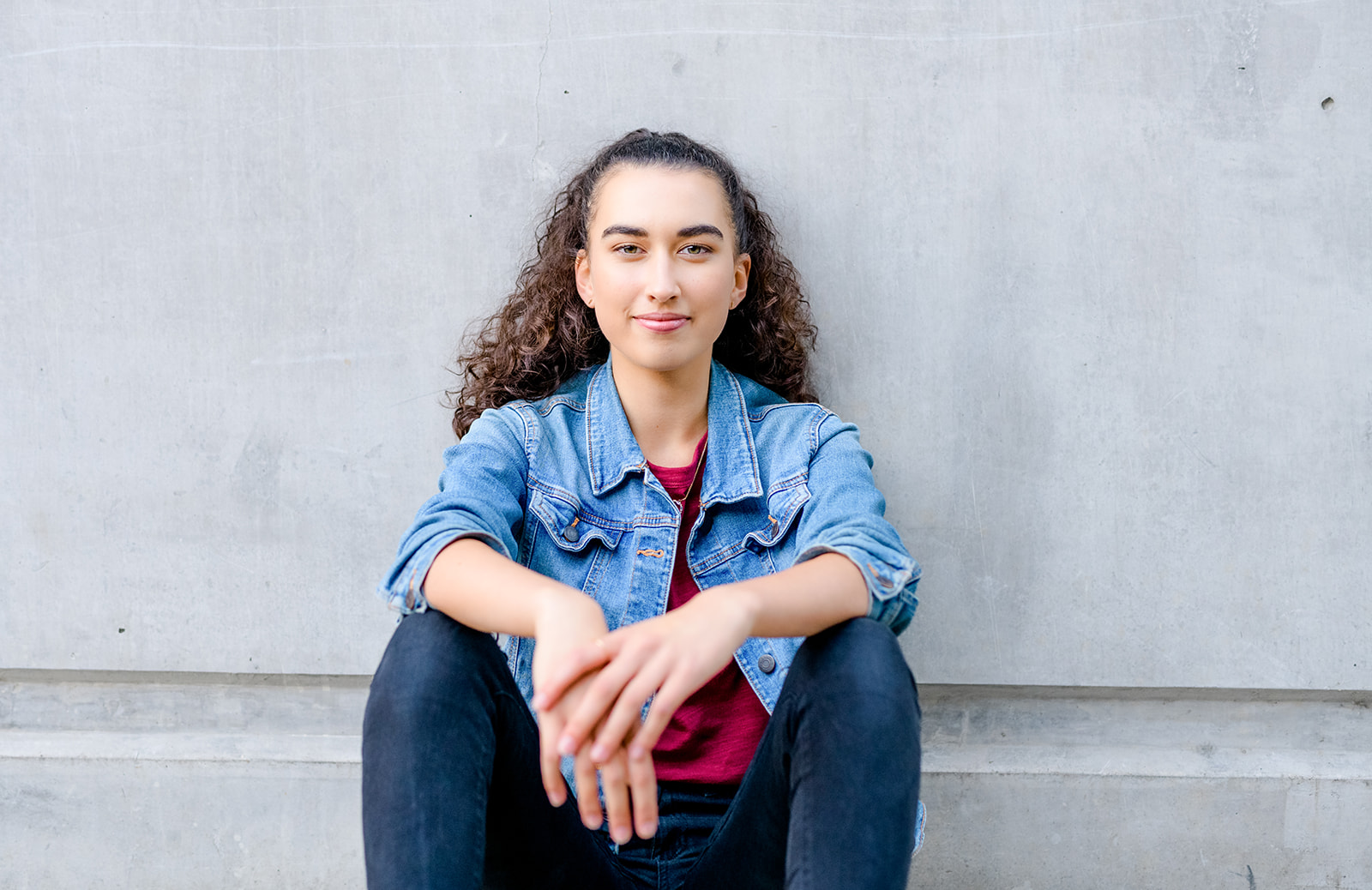 A high school senior sits against a concrete wall in a denim jacket and arms resting on her knees