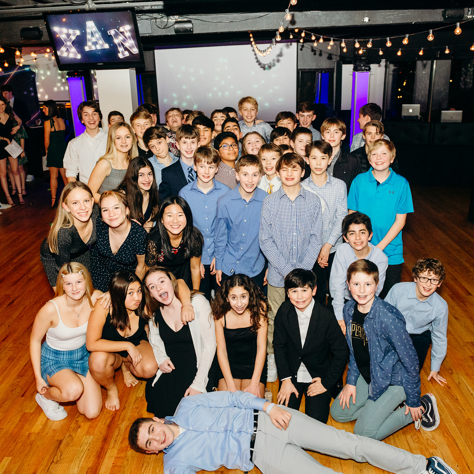 A large group of pre teen friends stand together on a dance floor during a Mitzvah