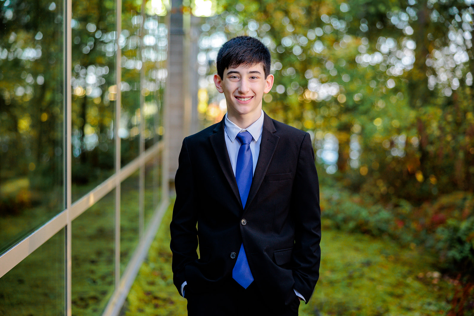 A teen boy stands in a garden along a glass wall building in a black suit and blue tie