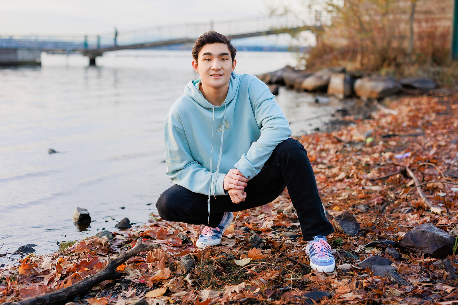 A high school senior boy sqauts on the edge of a river in fall wearing a blue hoodie