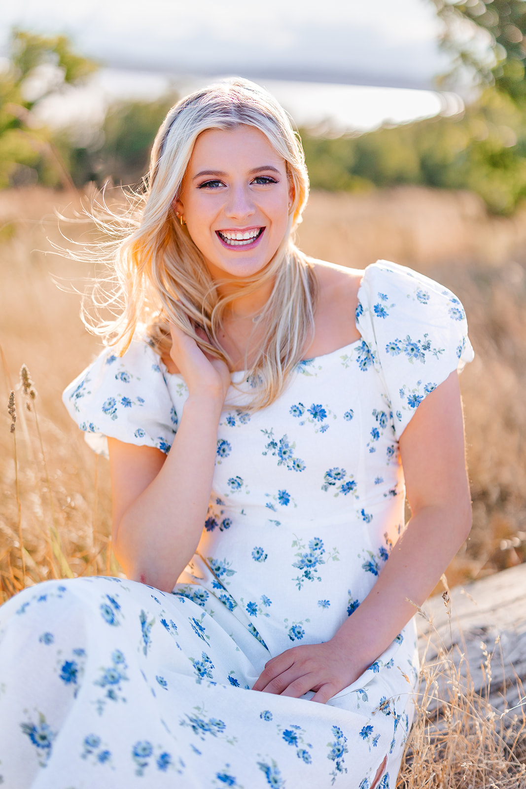 A high school senior in a white dress with blue flowers sits on a log in a windy field of golden grass at sunset after attending Bellevue high school Washington