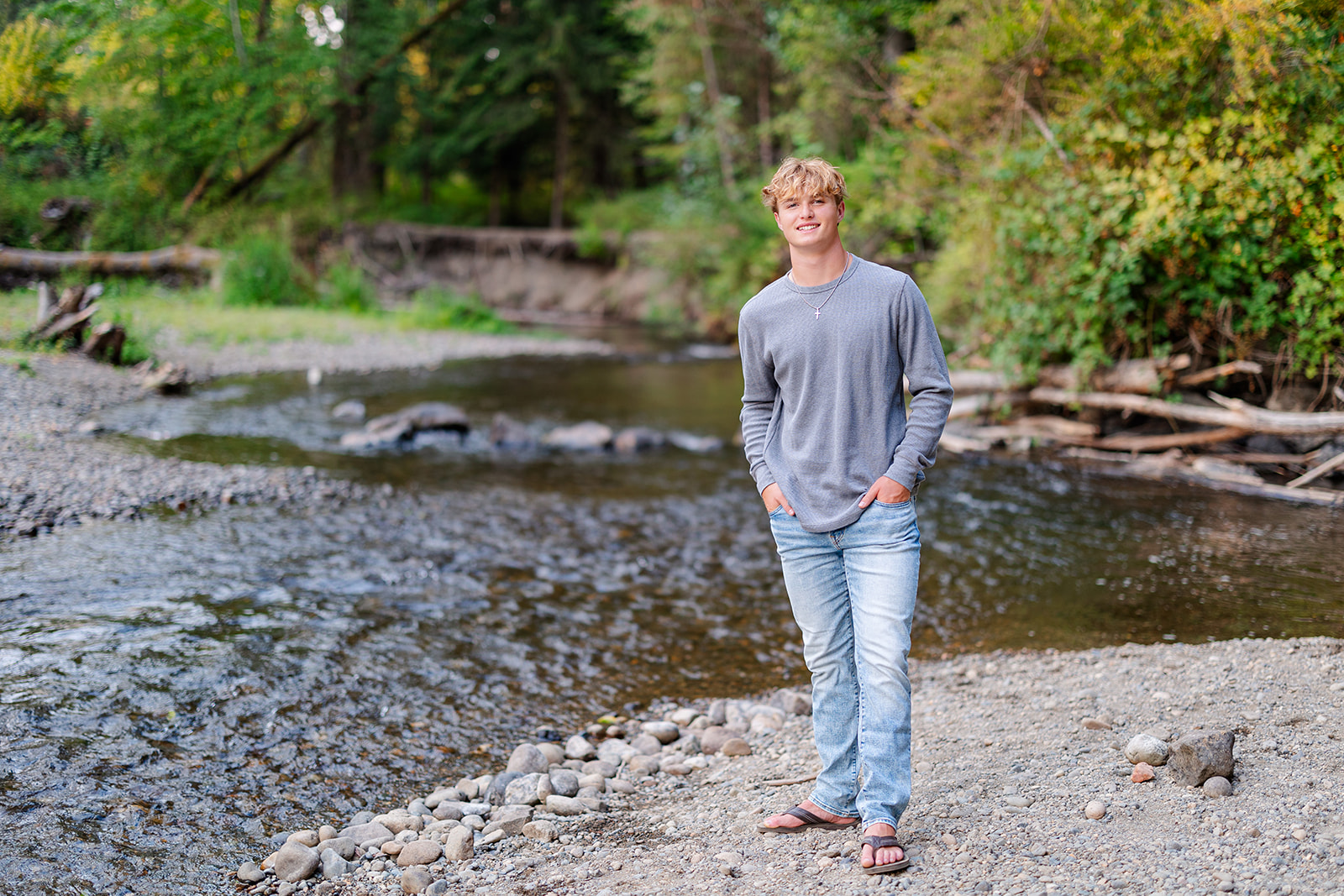 A high school senior in jeans and a grey shirt stands along a river in a park