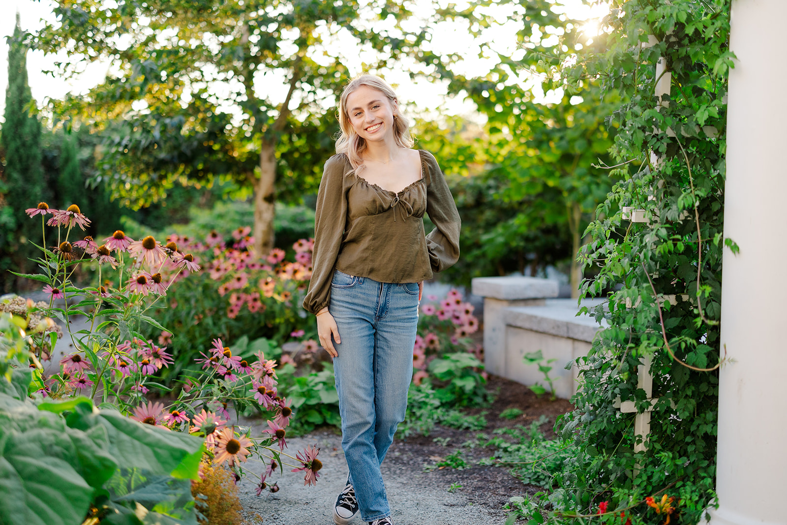 A high school senior walks through a lush garden at sunset in a green shirt and jeans