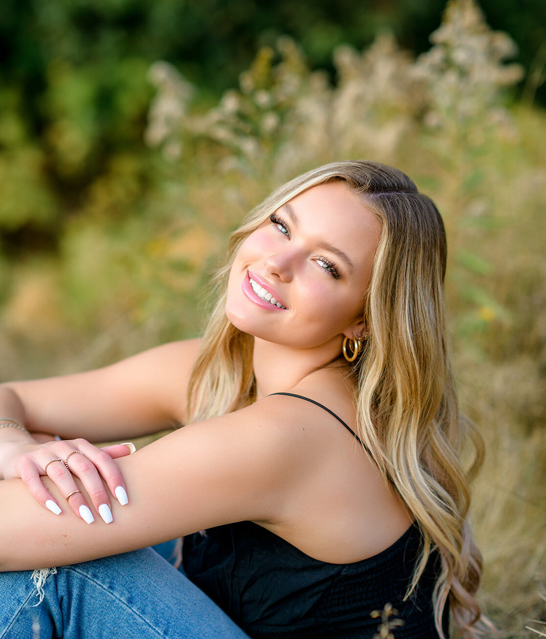 A high school senior in jeans and a black top sits in a field of tall grass smiling over her shoulder