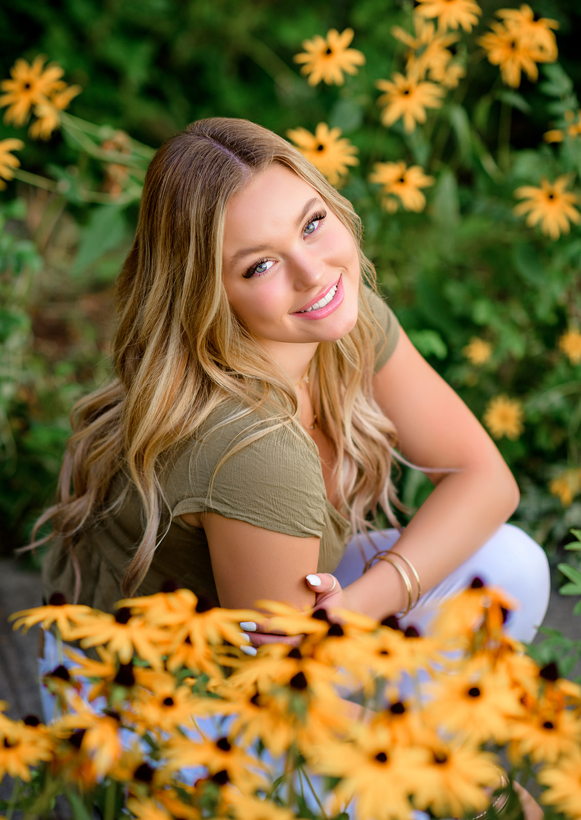 A high school senior with blonde hair sits among some yellow sunflowers in a green top after using Hair Salons Bellevue