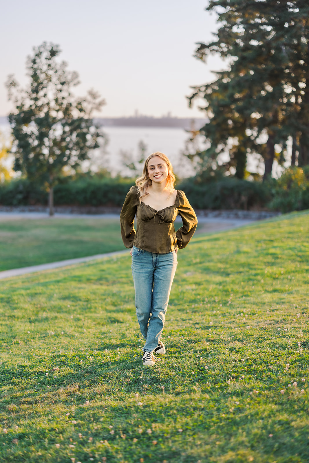 A high school senior in a green shirt and jeans walks through a park lawn at sunset