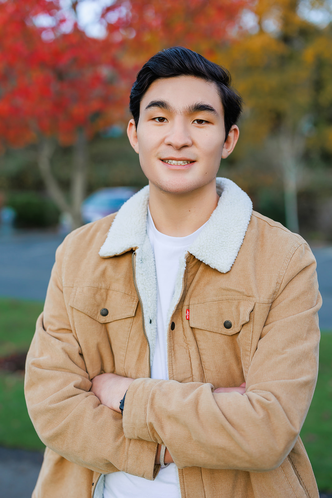 A high school senior in a tan denim jacket stands in a fall park with arms crossed