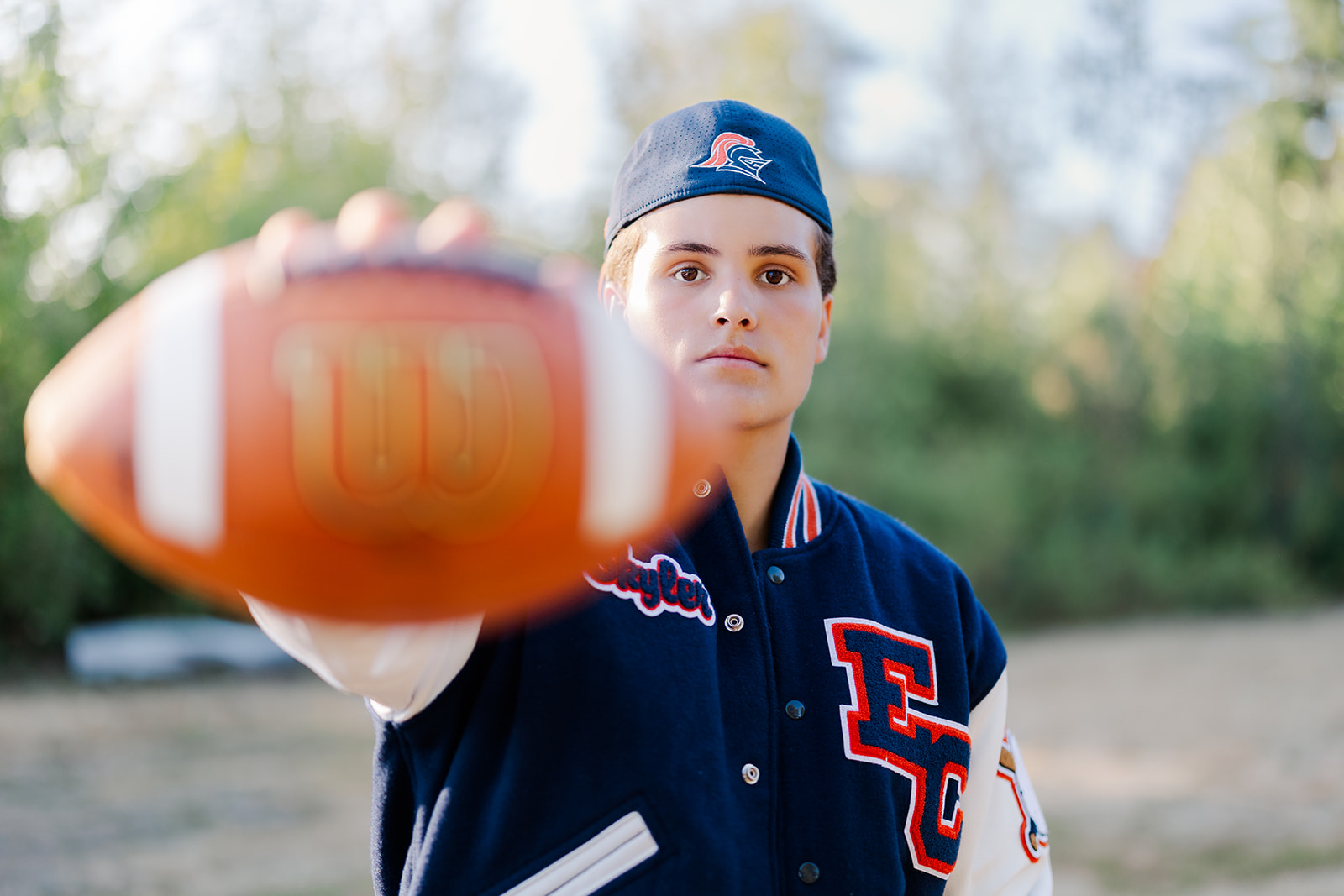 A high school senior stands in a park at sunset in a letterman jacket holding a football in front of him after finding high school senior volunteer opportunities seattle