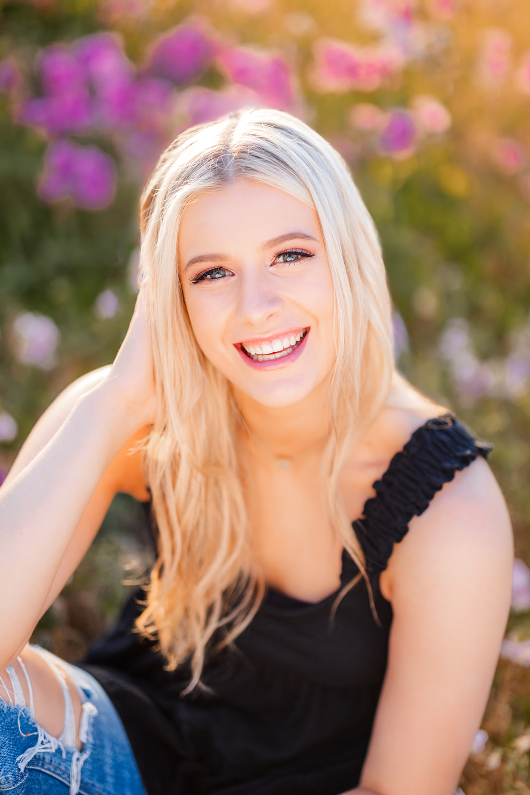 A high school senior sits in a field of wildflowers laughing at sunset in a black top