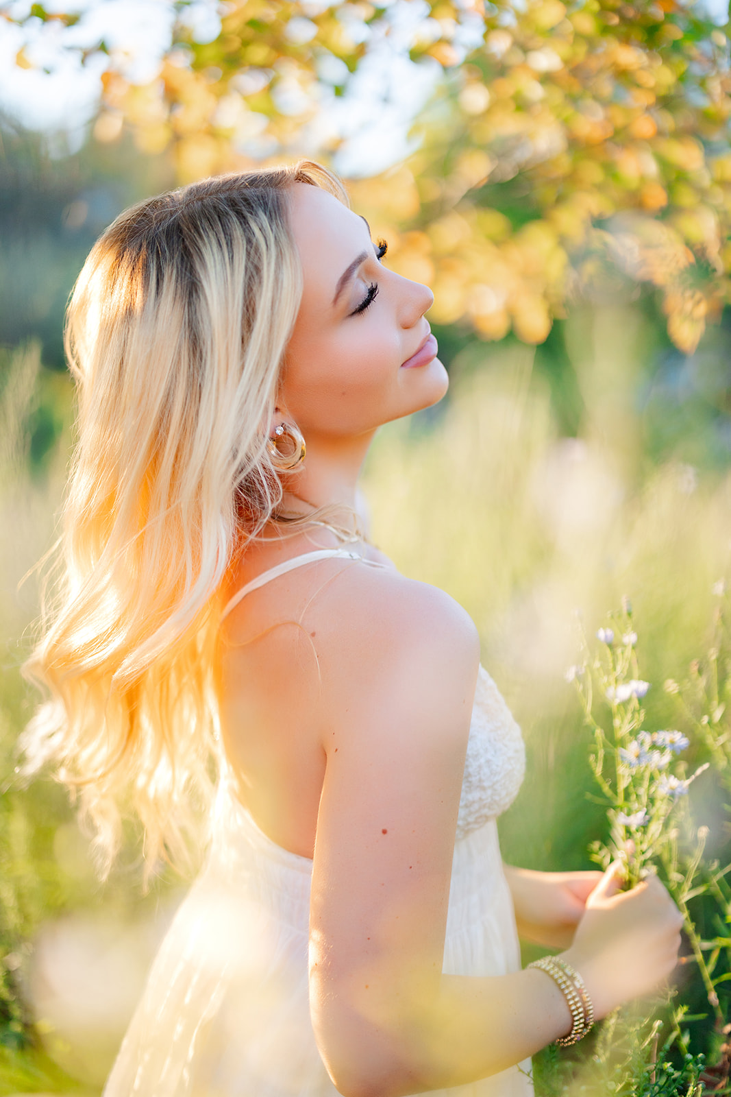 A high school senior picks wildflowers in a field in a white dress at sunset