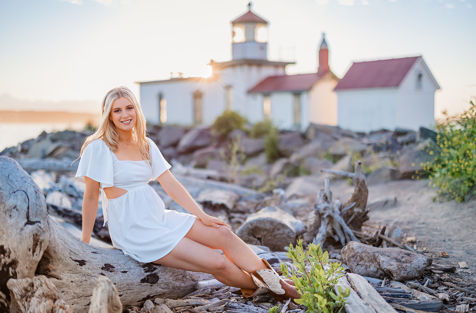 A high school senior sits on some driftwood on a beach at sunset in a white dress after using Makeup Artists Bellevue