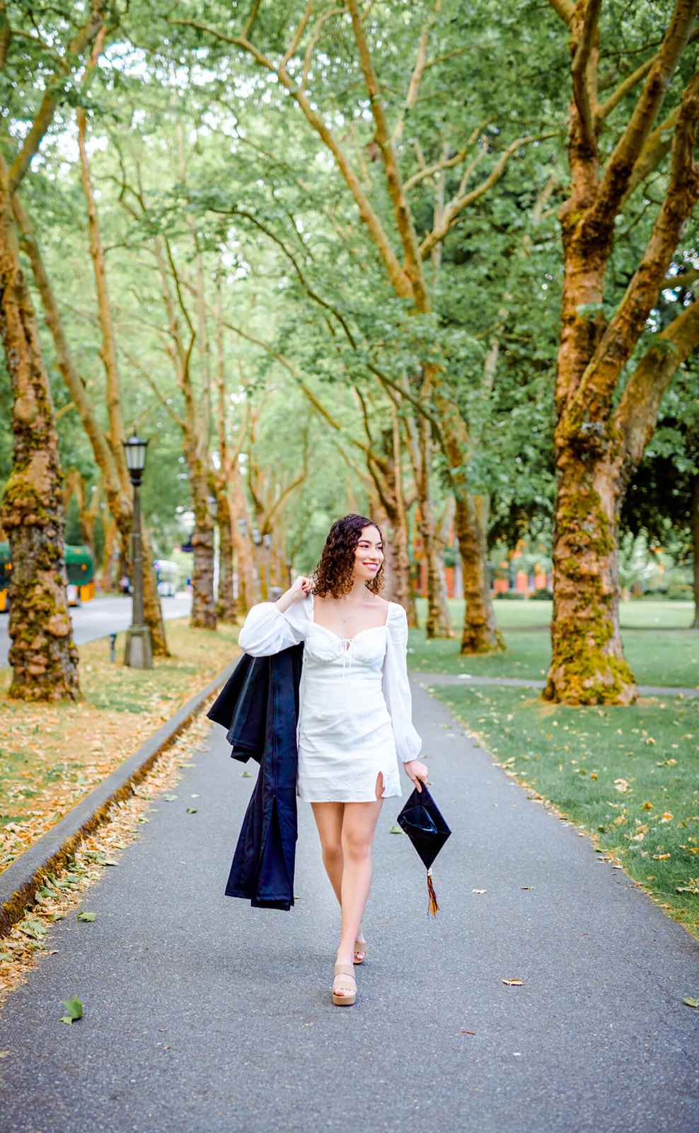 A high school senior in a white dress walks through a park sidewalk holding her cap and gown after graduating Mercer Island High School