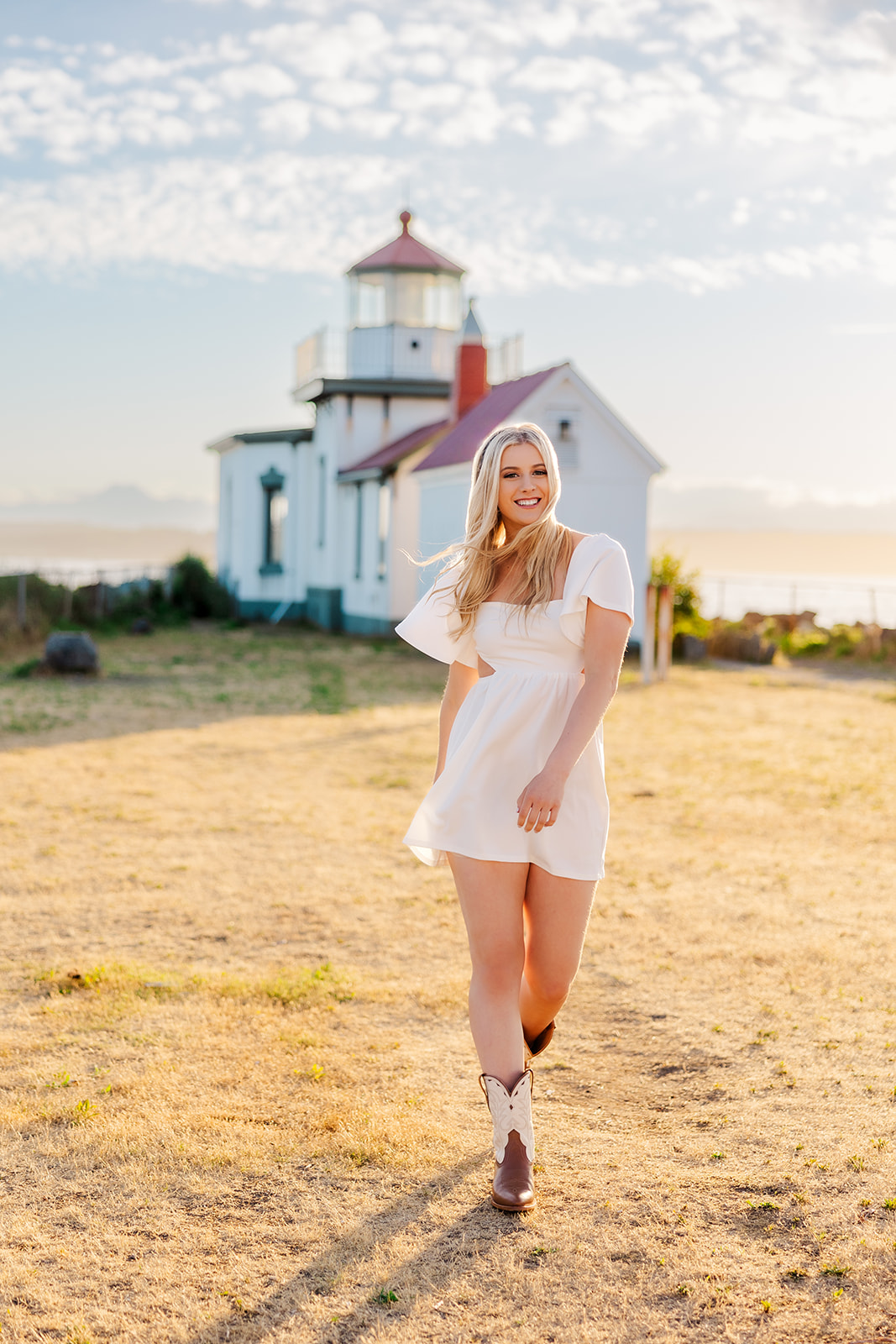 A high school senior in a white dress walks away from a lighthouse at sunset