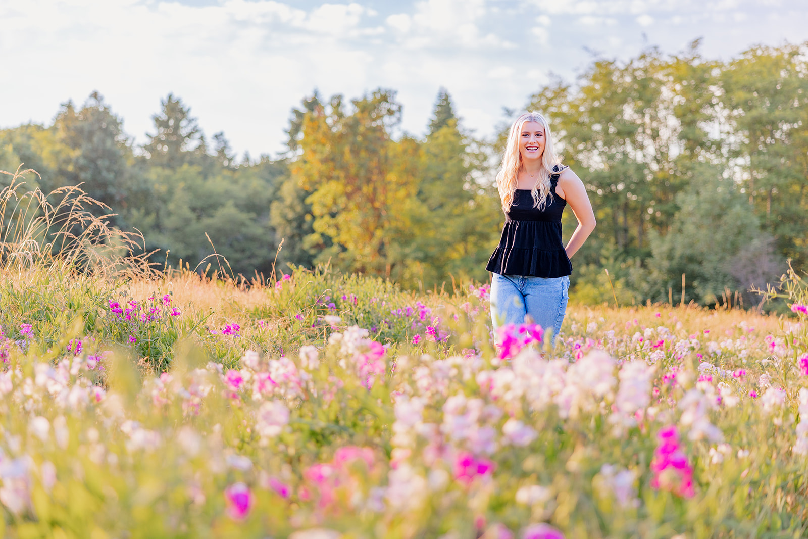 A high school senior in jeans and a black top walks through a field of wildflowers at sunset after some Nail Salons Bellevue