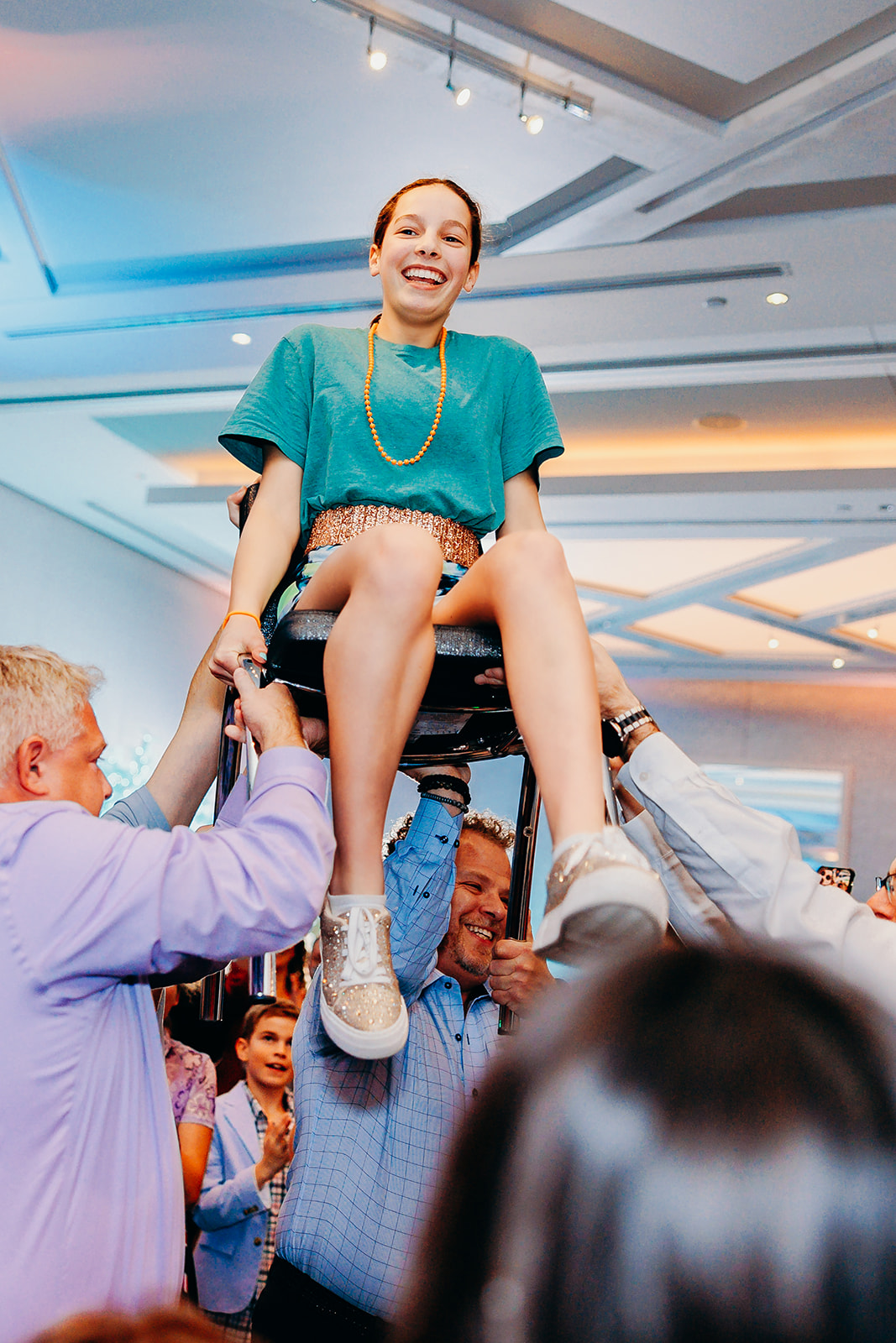 A girl in a teal dress sits on a chair being lifted high in the air on a dance floor during her Mitzvah while using Party Entertainment Seattle