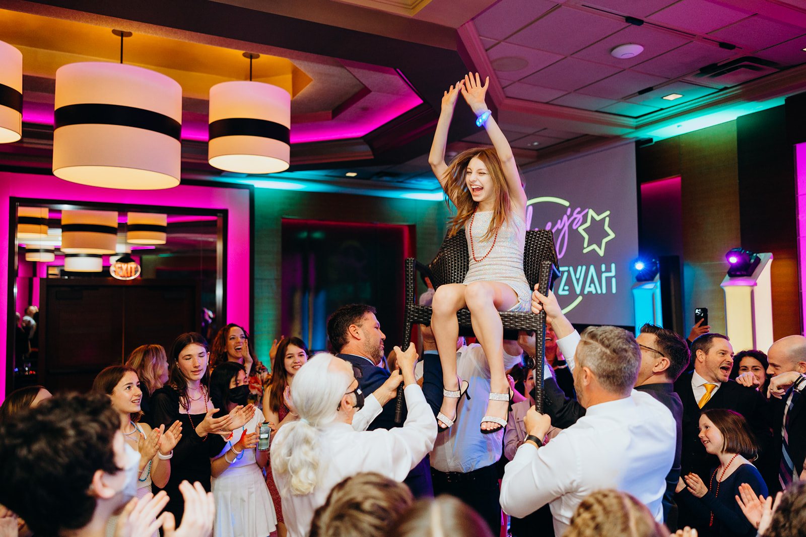 A girl in a white dress celebrates her Mitzvah sitting on a chair while being lifted in the air with help from some Party Entertainment Seattle