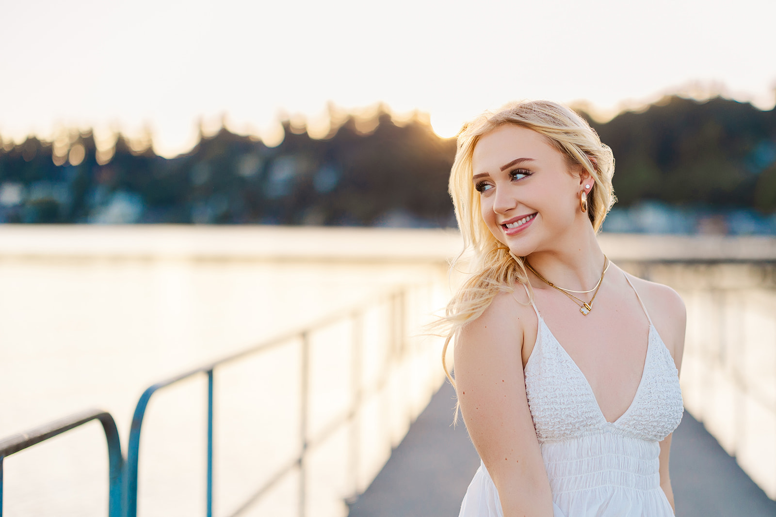 A high school senior in a white dress smiles over her shoulder while walking on a dock at sunset