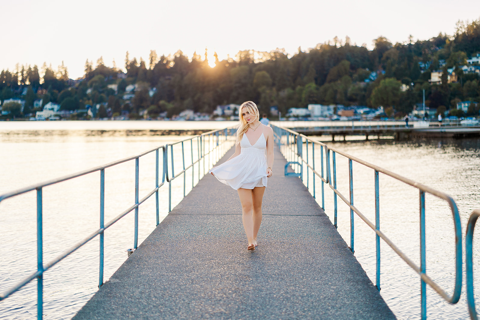 A high school senior walks barefoot on a dock in a white dress at sunset before visiting Prom Dress Shops In Seattle