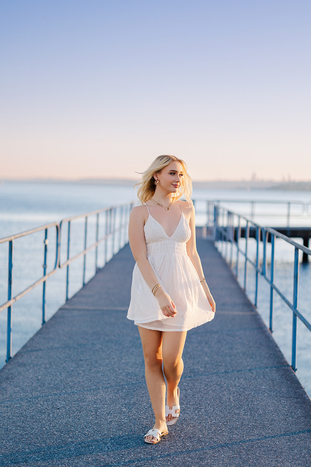 A high school senior in a white dress walks on a windy day up a dock on a river at sunset after visiting Prom Dress Shops In Seattle