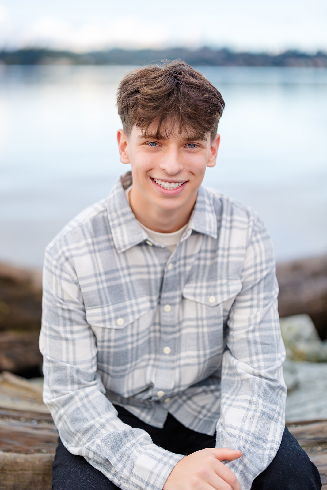 A high school senior sits on a piece of driftwood on waters edge in a grey plaid shirt