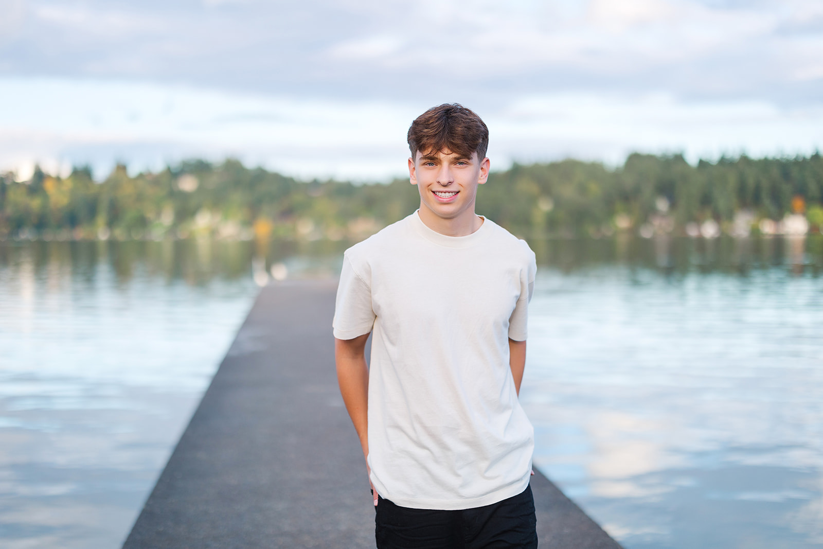 A high school senior stands on a dock with his hands in his back pockets smiling