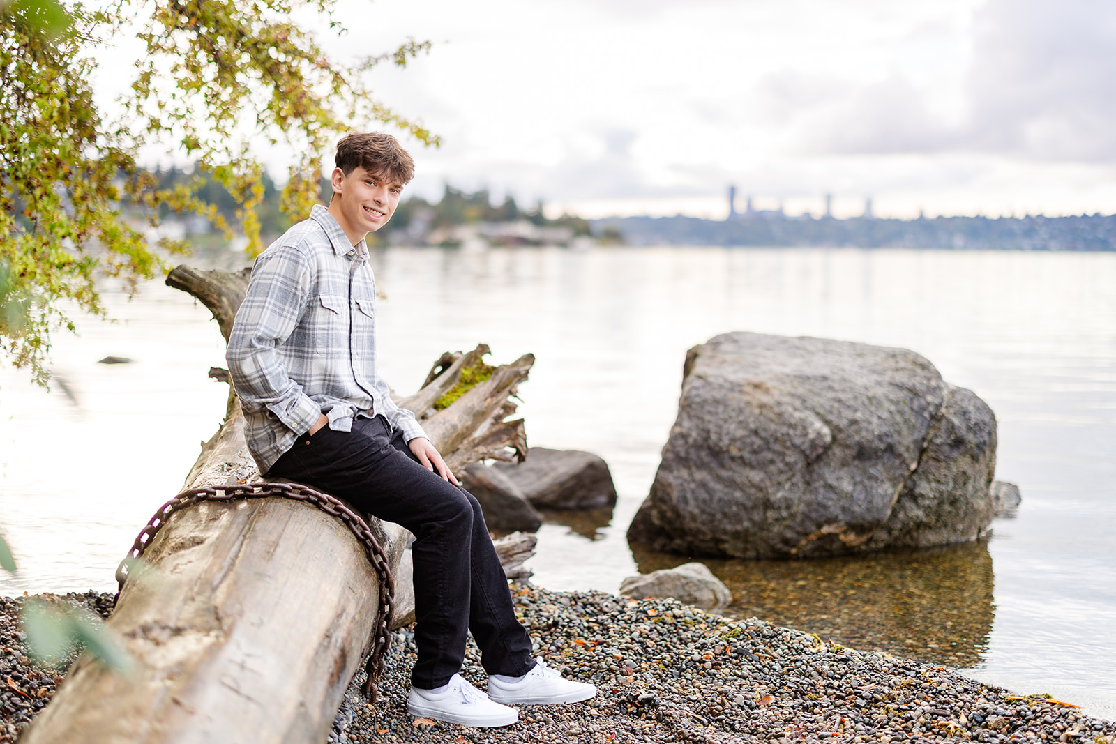 A high school senior sits on a fallen tree on a river in a plaid shirt and black pants before some SAT Prep Bellevue