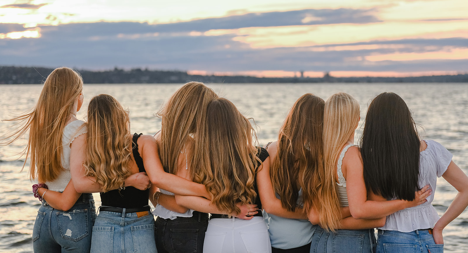 A group of girlfriends stand on a dock with arms around each other while watching the sunset over the water