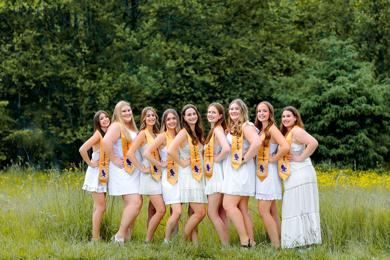 A group of friends in white dresses and gold stoles stand in a field of wildflowers with hands on their hips before visiting Tanning Salons in Bellevue