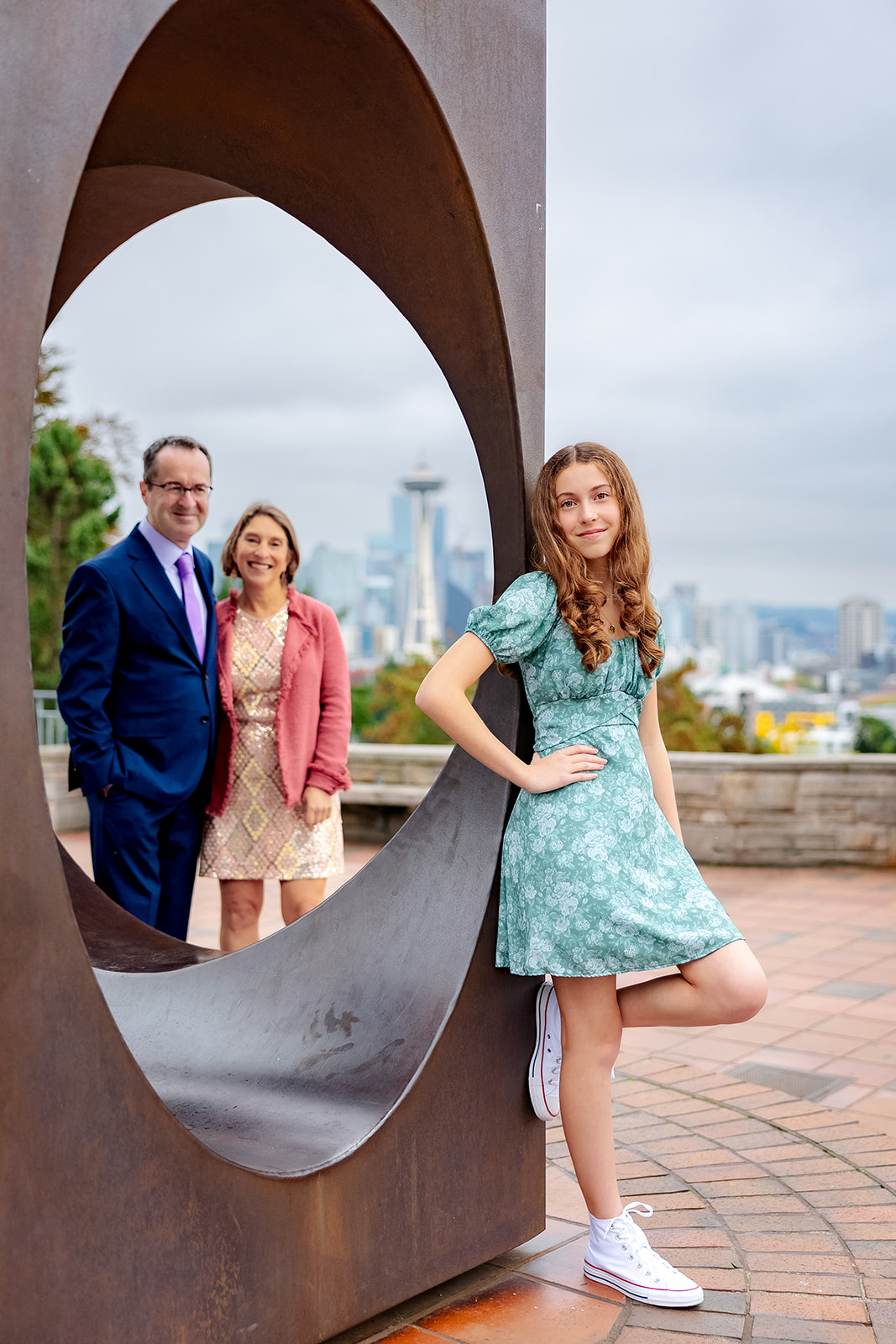 A teenager leans against a park sculpture with mom and dad looking on before her Bat Mitzvah