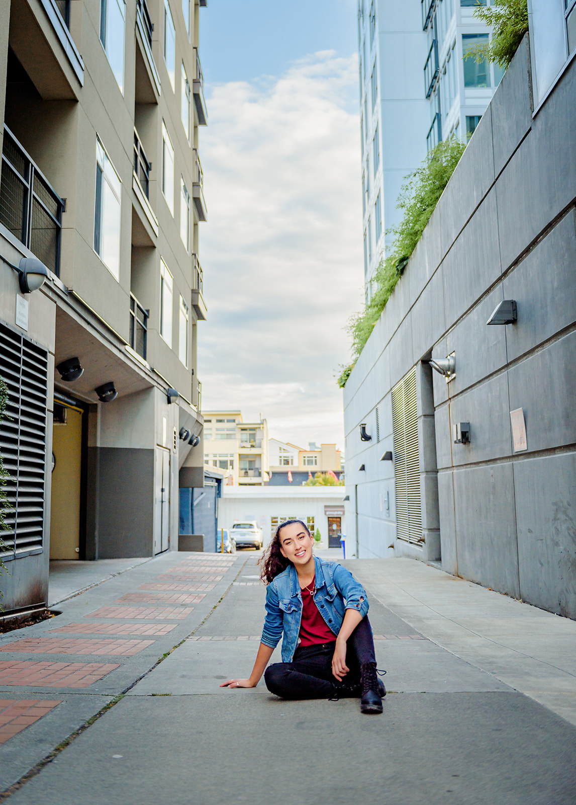 A high school senior sits in an alley in a jean jacket and red top