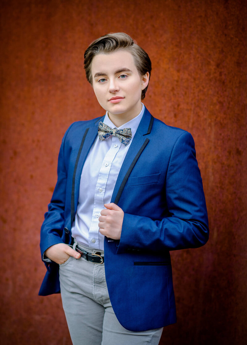 A teenager stands in a studio holding the lapels on their blue tuxedo jacket