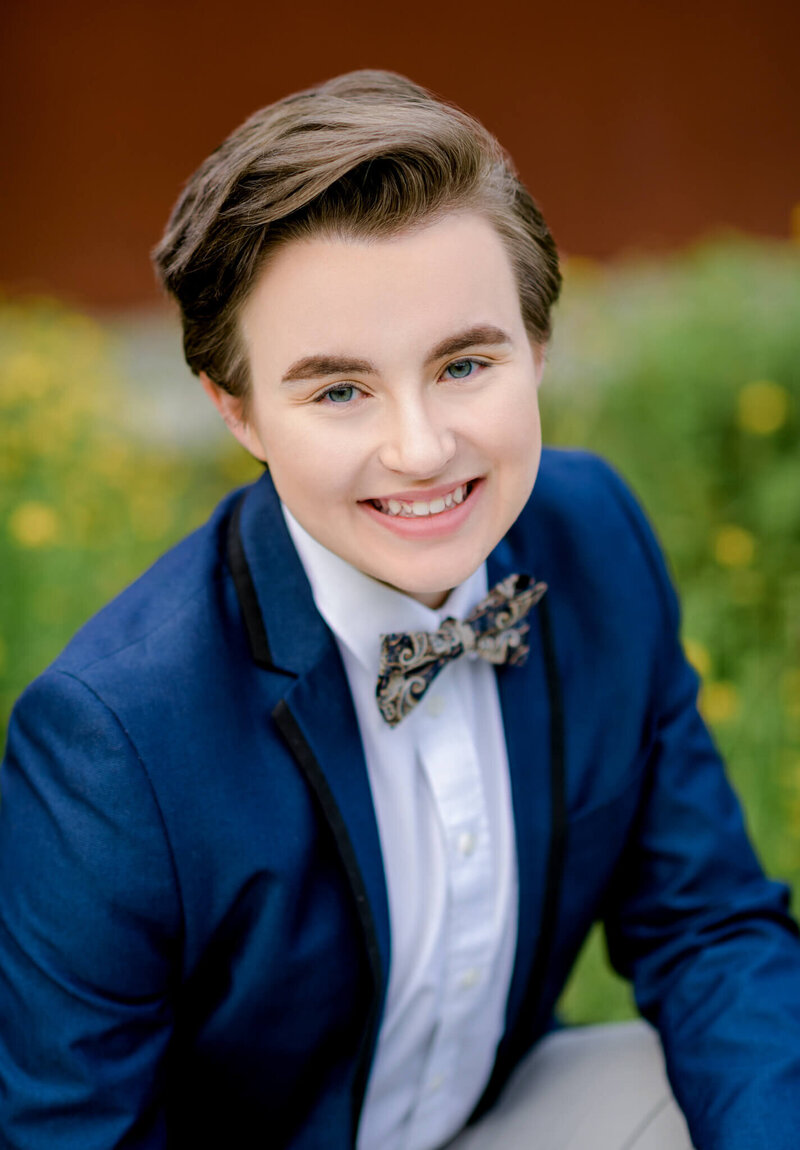 A teenager smiles while sitting in a pattern bowtie and blue suit from a tux shop bellevue