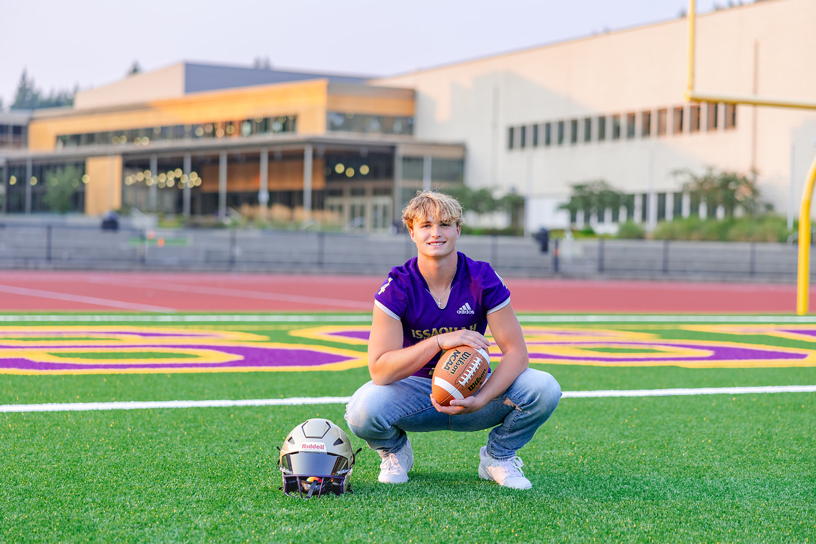 A high school senior in a football jersey holds a football while squatting on a football field