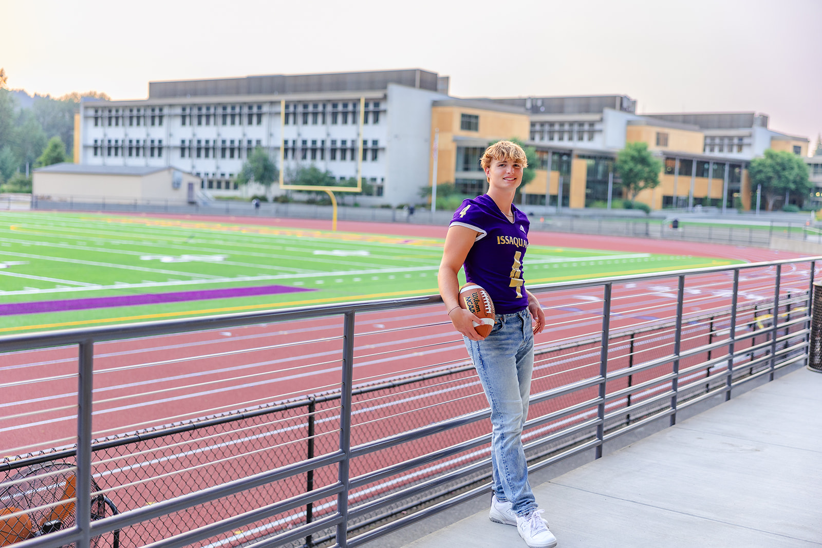 A high school senior leans on a railing in the football stadium stands holding a football before Applying To Bellevue College