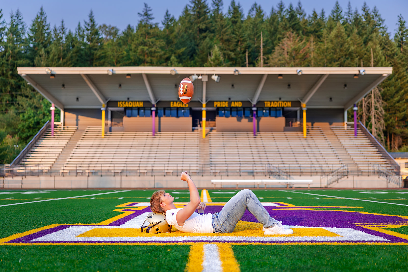 A high school senior football player lays on the painted logo at the 50 yard line throwing a football above his head before Applying To Bellevue College