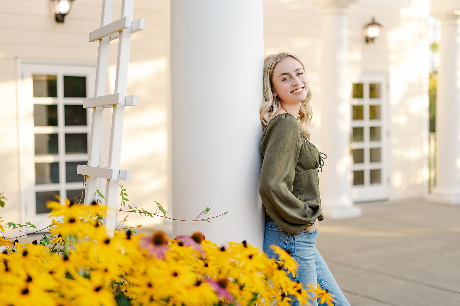 A happy high school senior leans on a white column in a garden with hands in her pockets after visiting Mercer Island Hair Salons