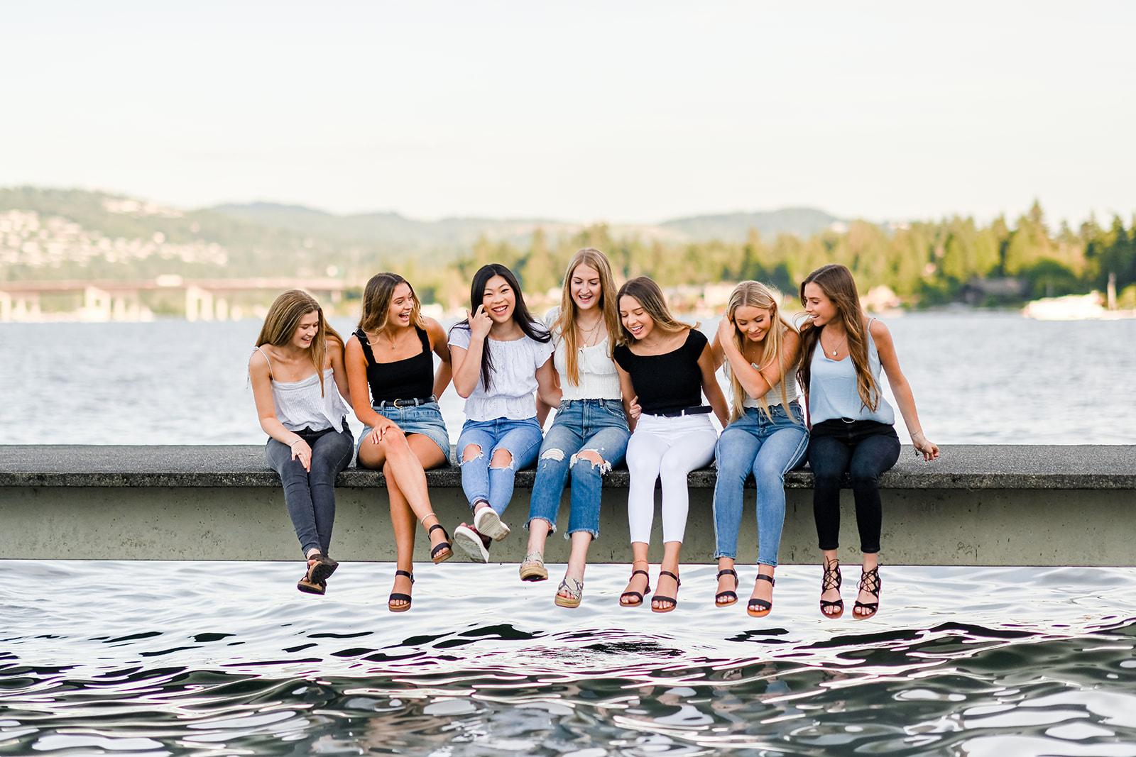 A group of happy high school senior girls sit on a concrete dock laughing and looking down into the water