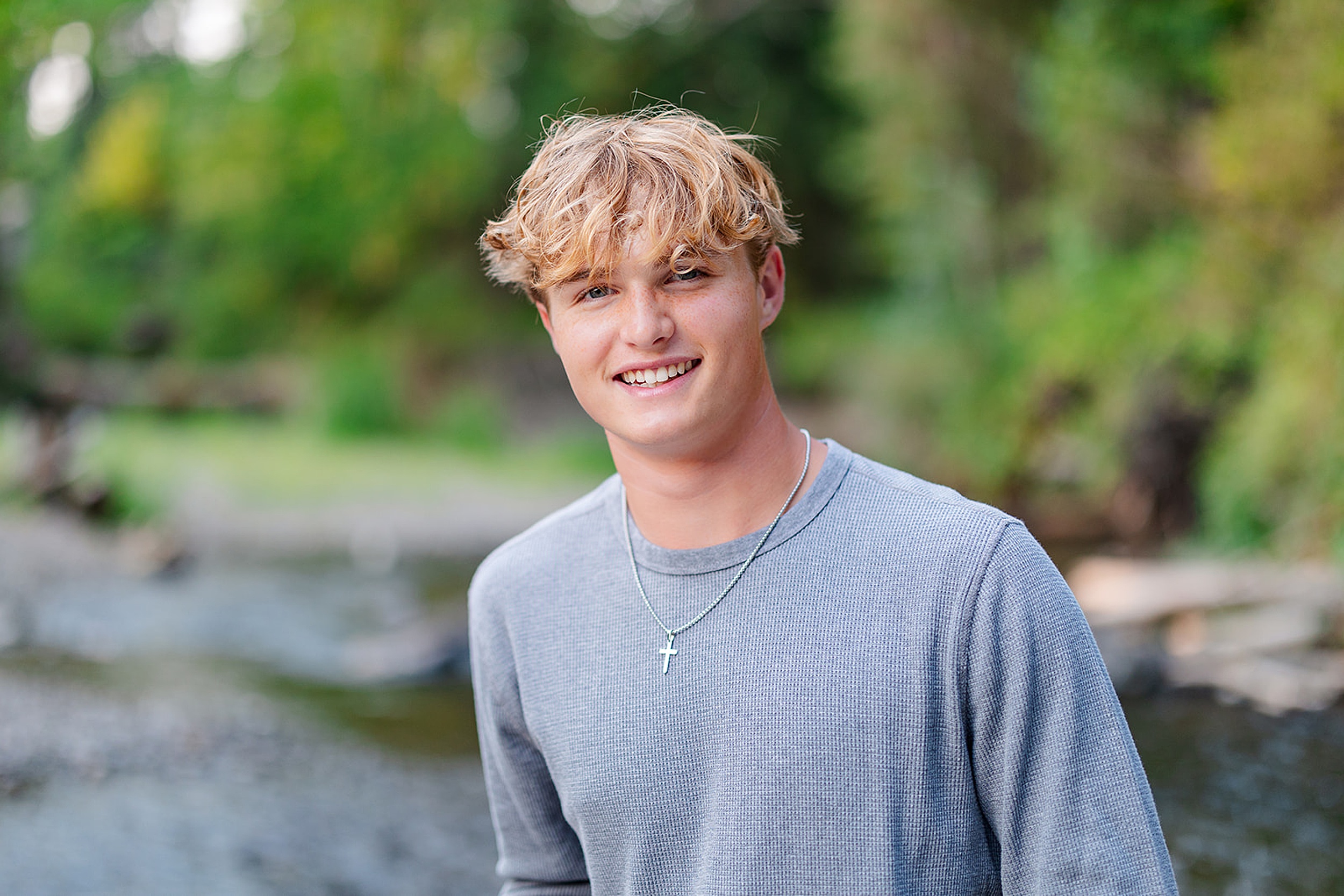 A high school senior in a grey long sleeve shirt stands by a flowing creek at sunset