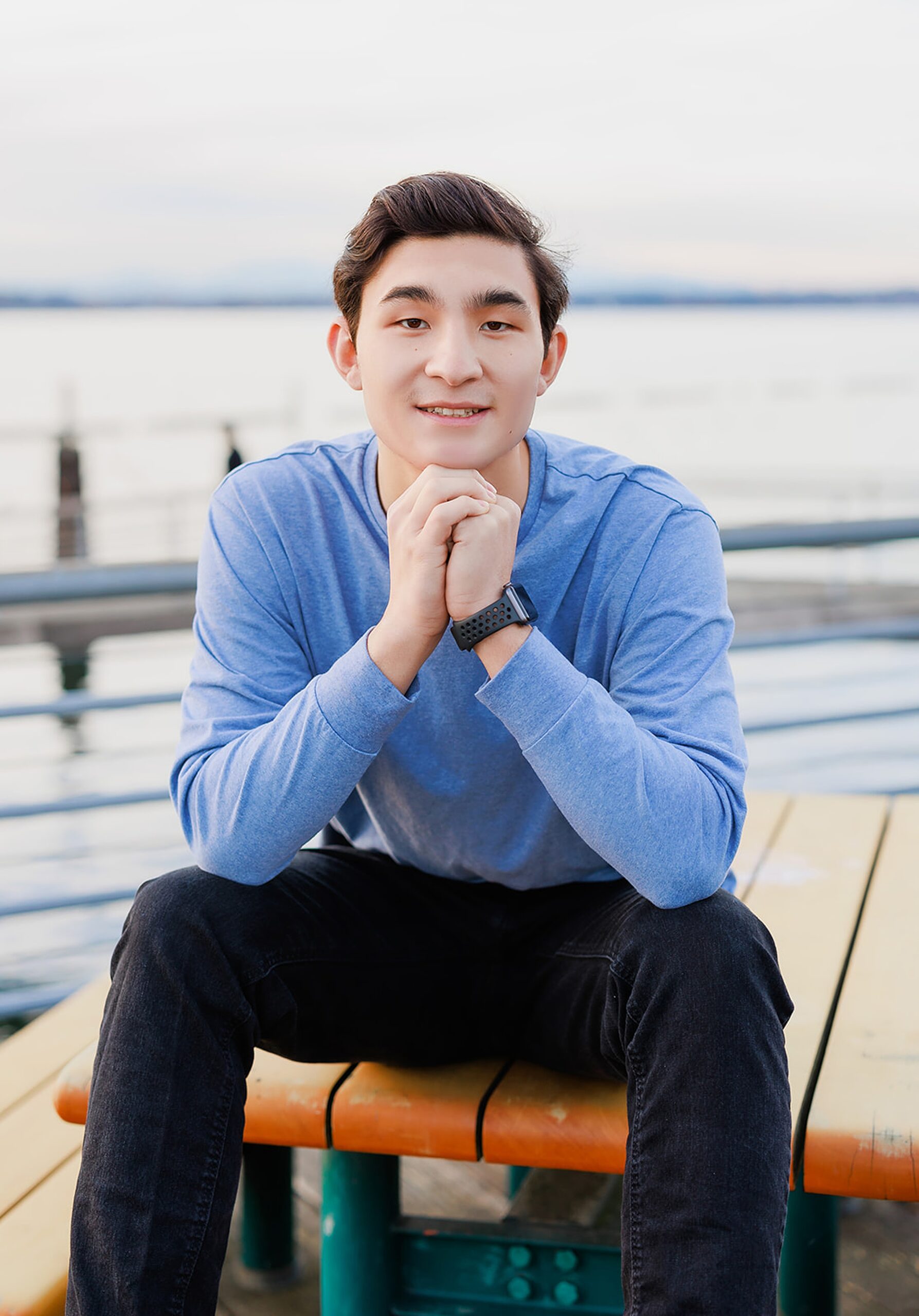 A high school senior sits on a picnic bench on a dock in a blue shirt resting his head on his hands after attending Mercer Island Private Schools