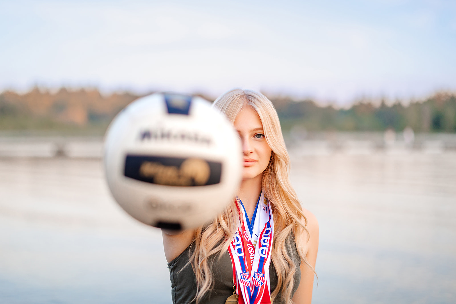 A high school senior with blonde hair stands on the edge of a river at sunset holding out a volleyball