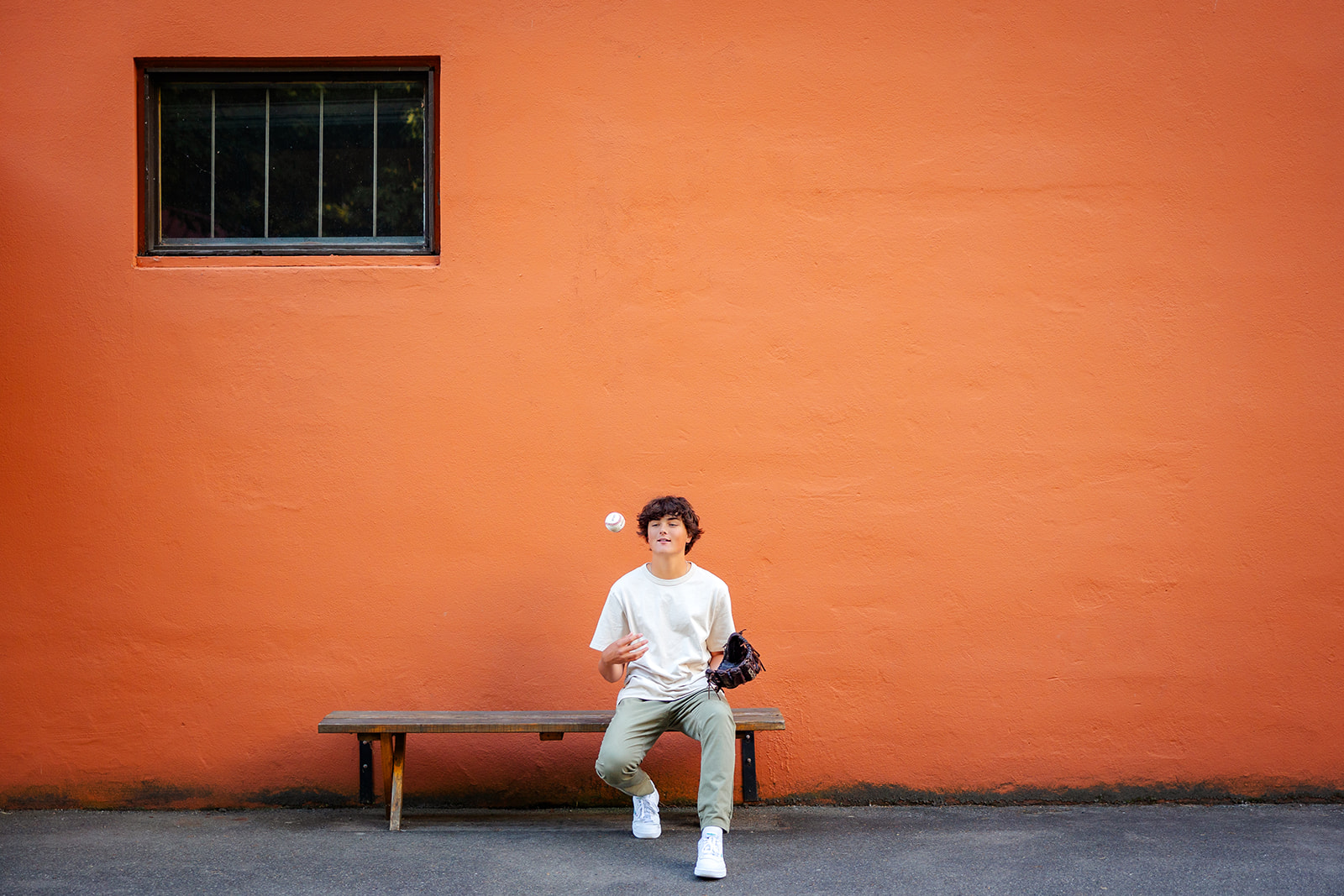 A high school senior sits on a bench against an orange wall tossing a baseball after playing Mercer Island Sports