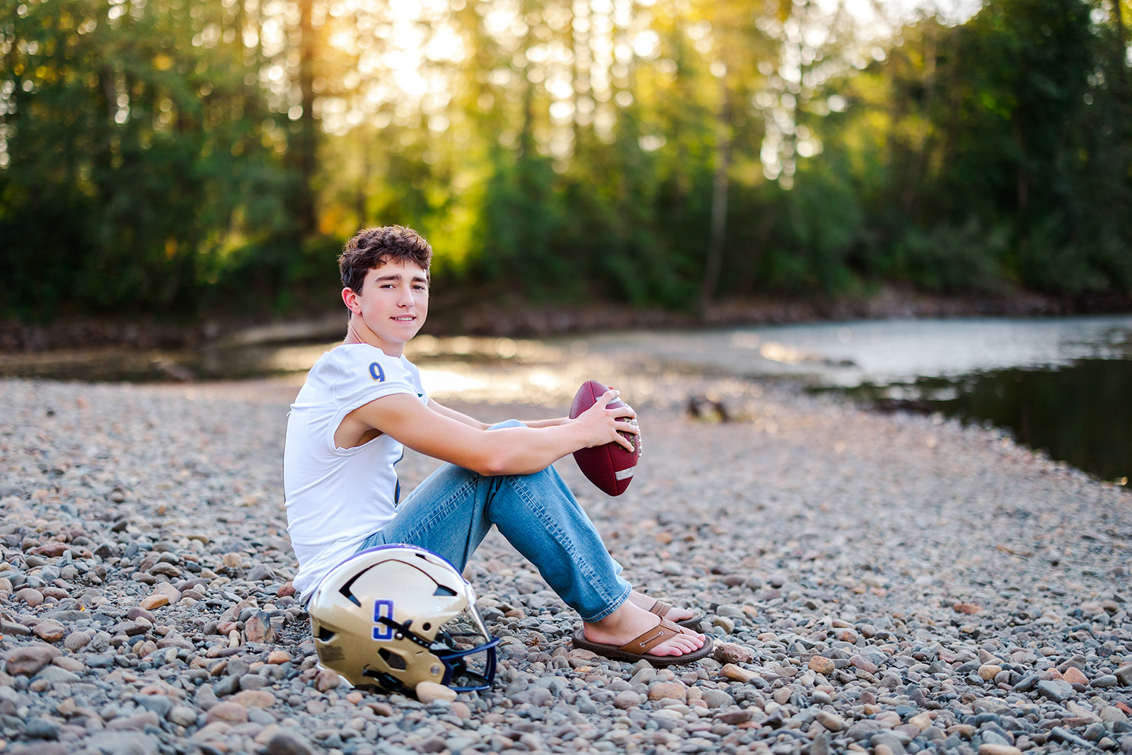 A high school senior football player sits on a rocky river bank in jeans and a jersey holding a football at sunset after playing Mercer Island Sports