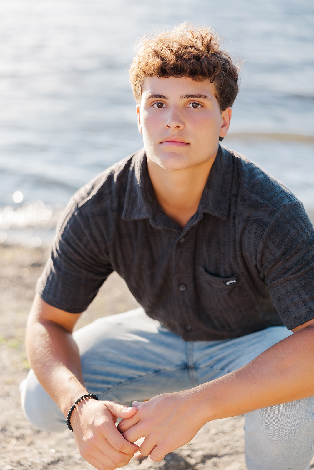 A high school senior squats on a beach in a button down shirt and jeans at sunset