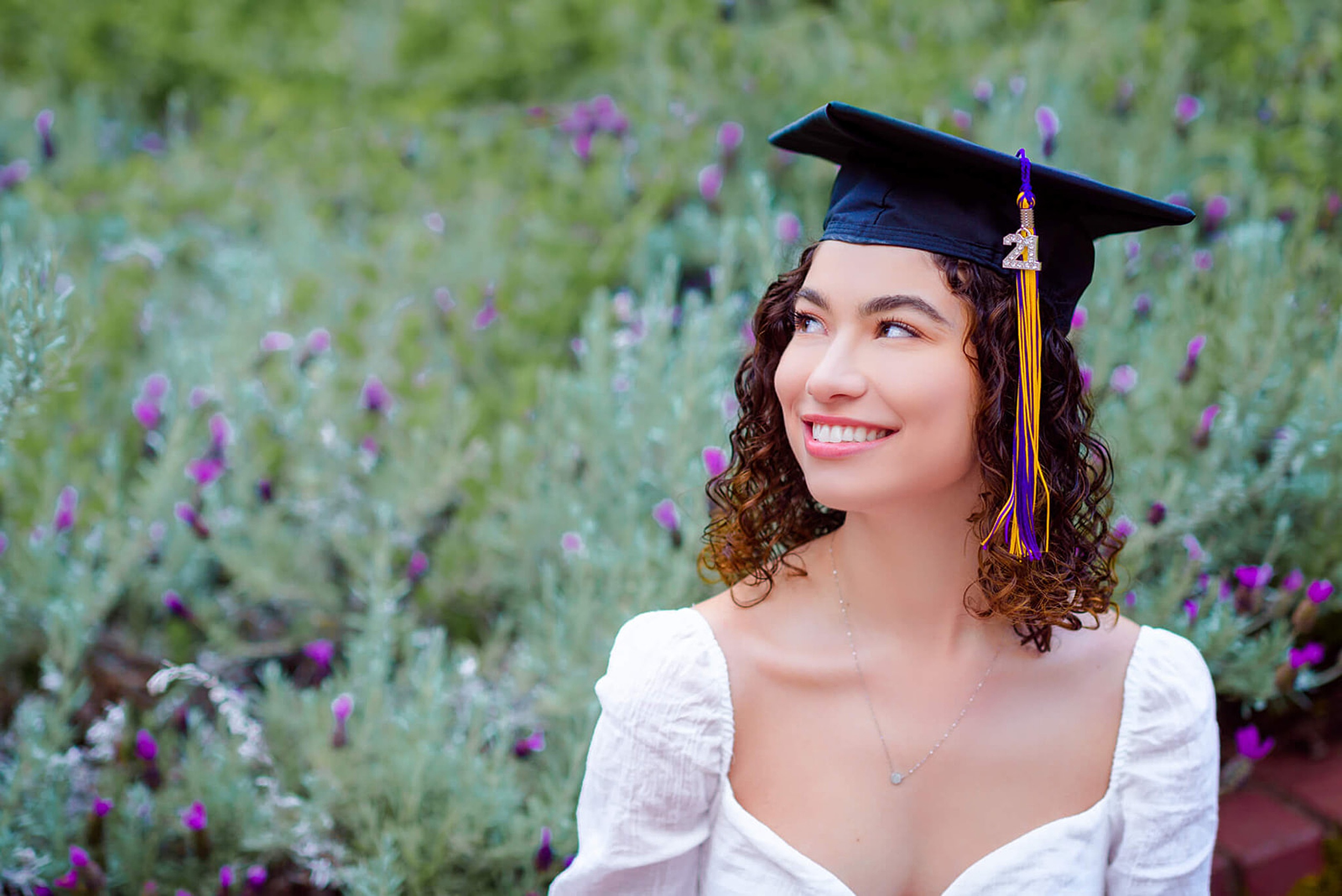 A graduate smiles big while sitting in a flower garden wearing a white dress and graduation cap