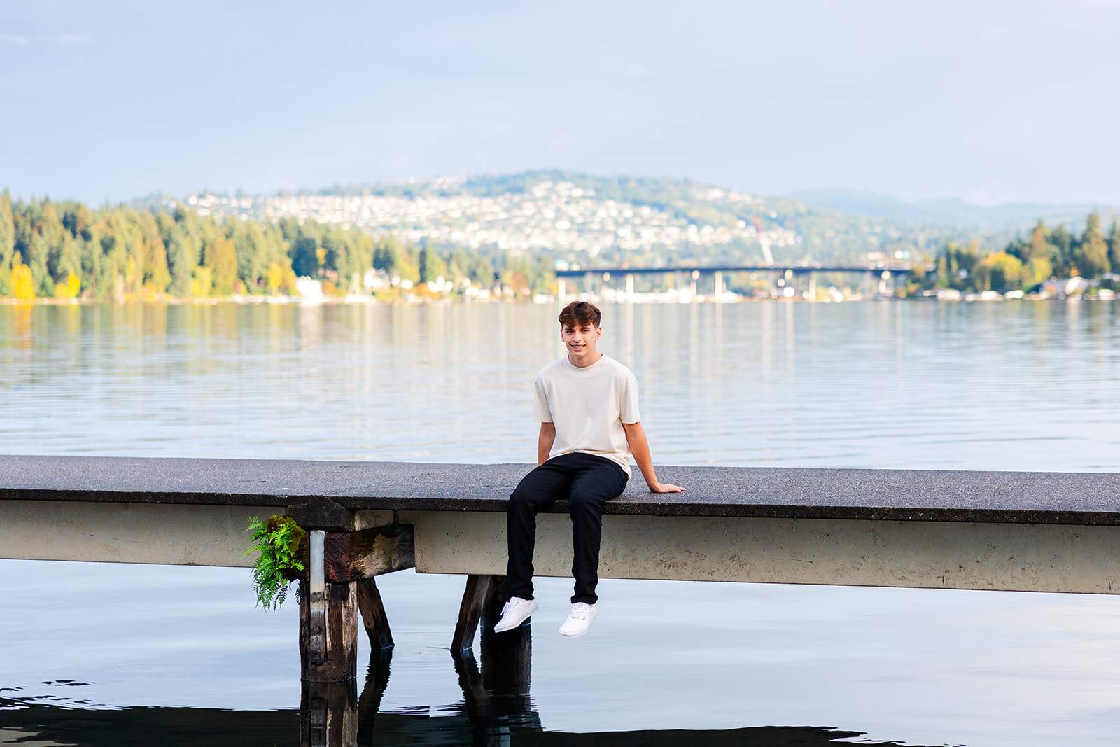 A high school senior sits on a dock in black pants and white shirt smiling after attending Torah Day School Of Seattle