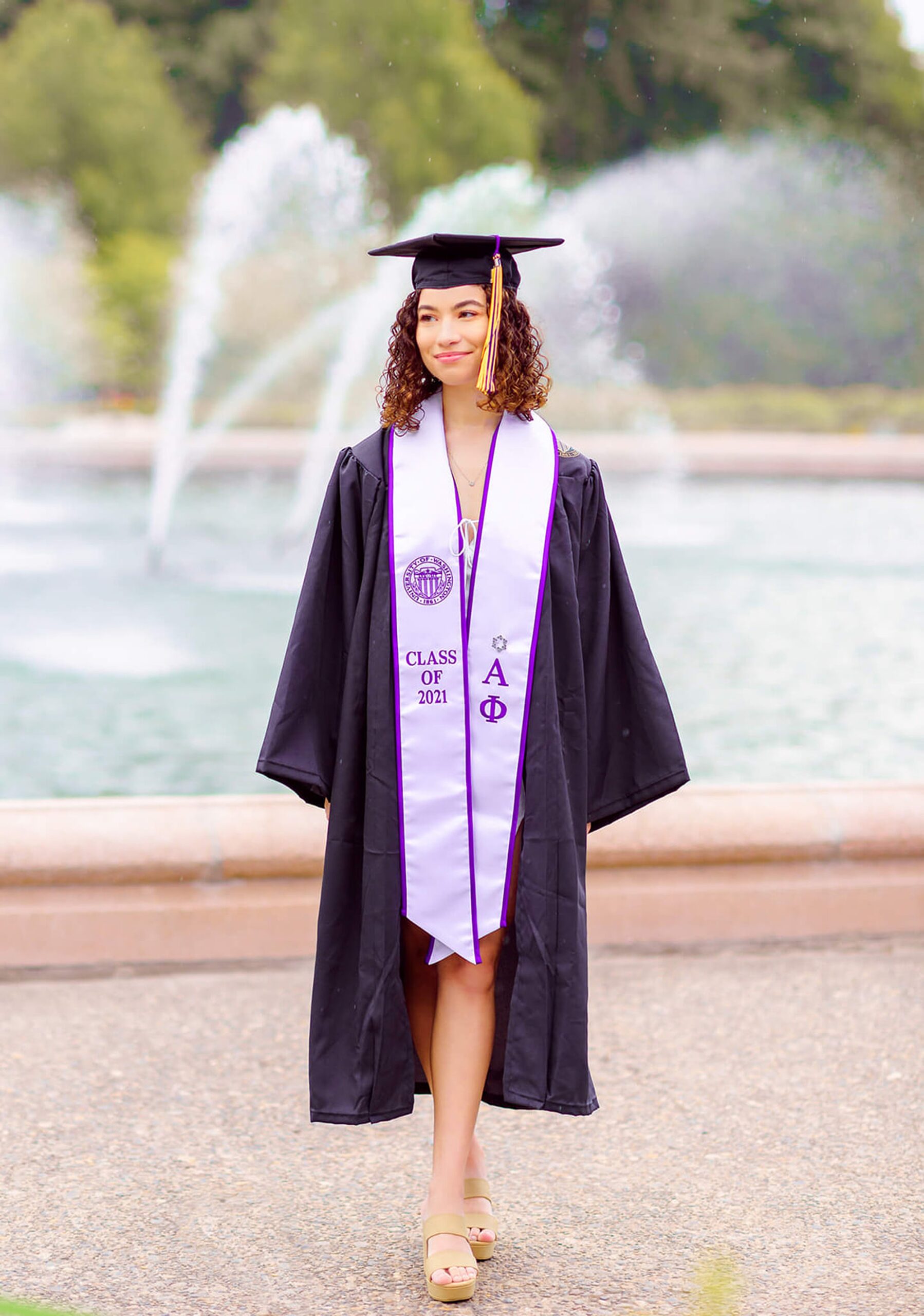 A graduate in a cap, gown and stole walks on a sidewalk in front of a fountain