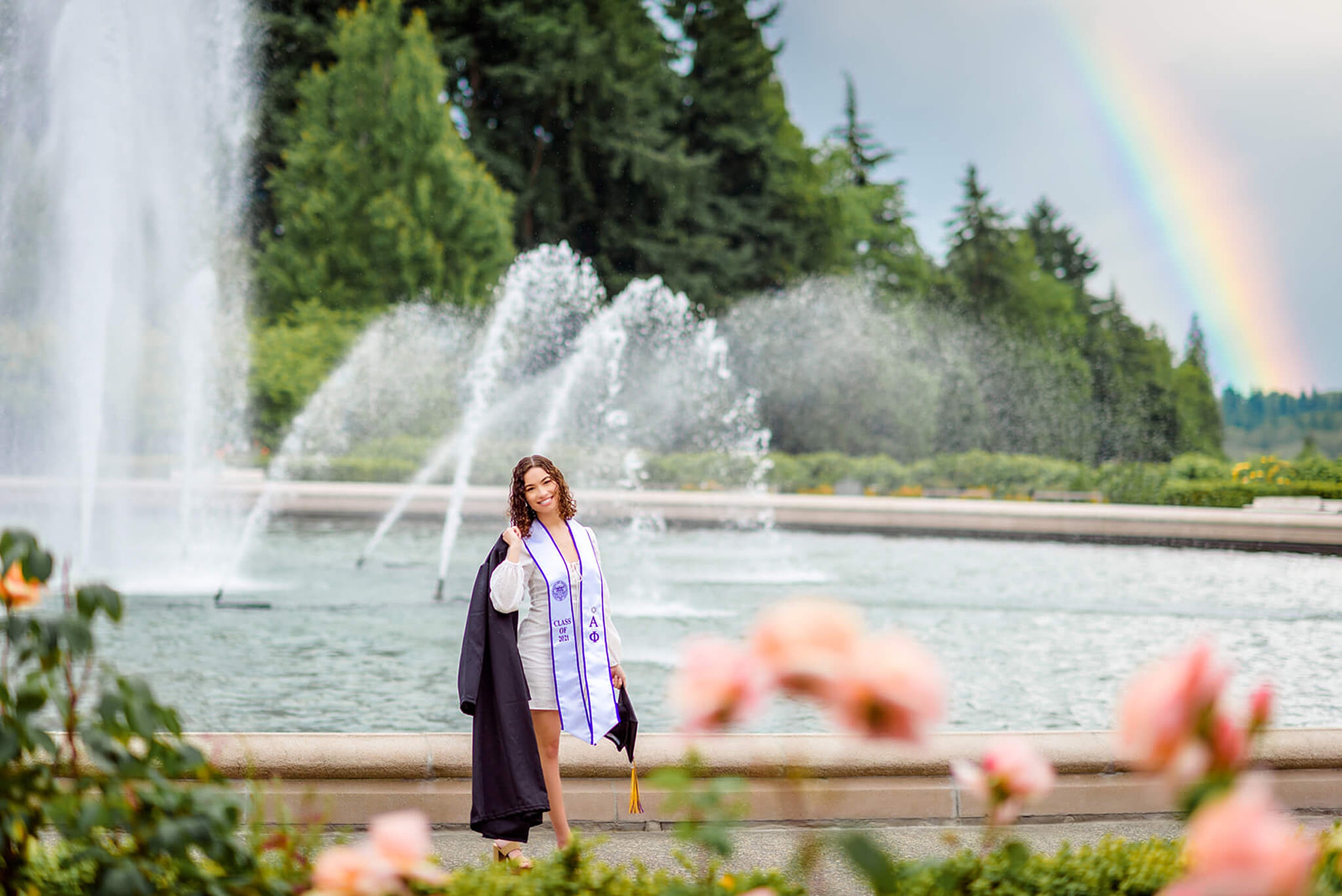 A graduate walks by a fountain wearing a white dress and stole while carrying her cap and gown after getting Tutoring Mercer Island