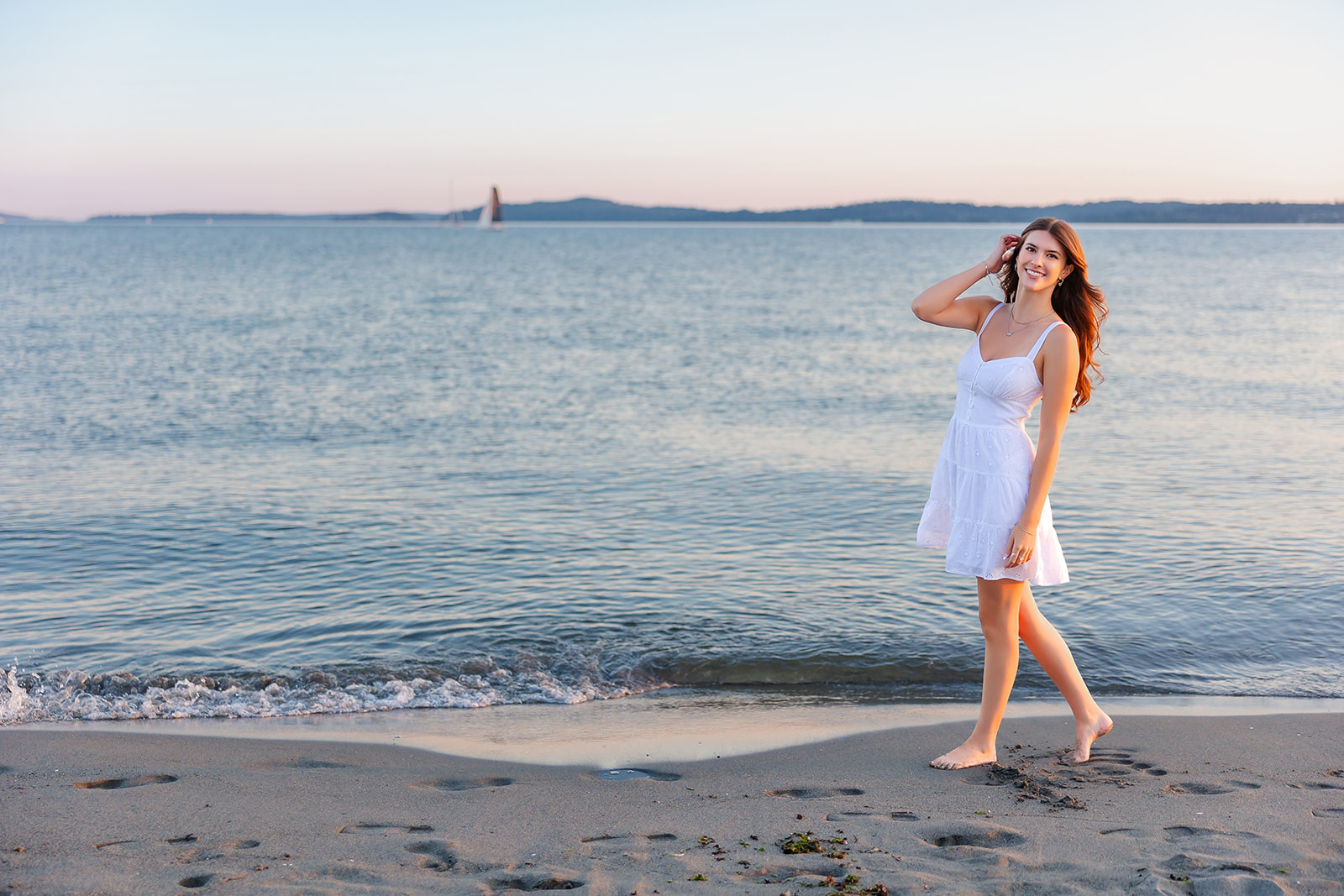 A high school senior walks barefoot on a beach in a white dress at sunset