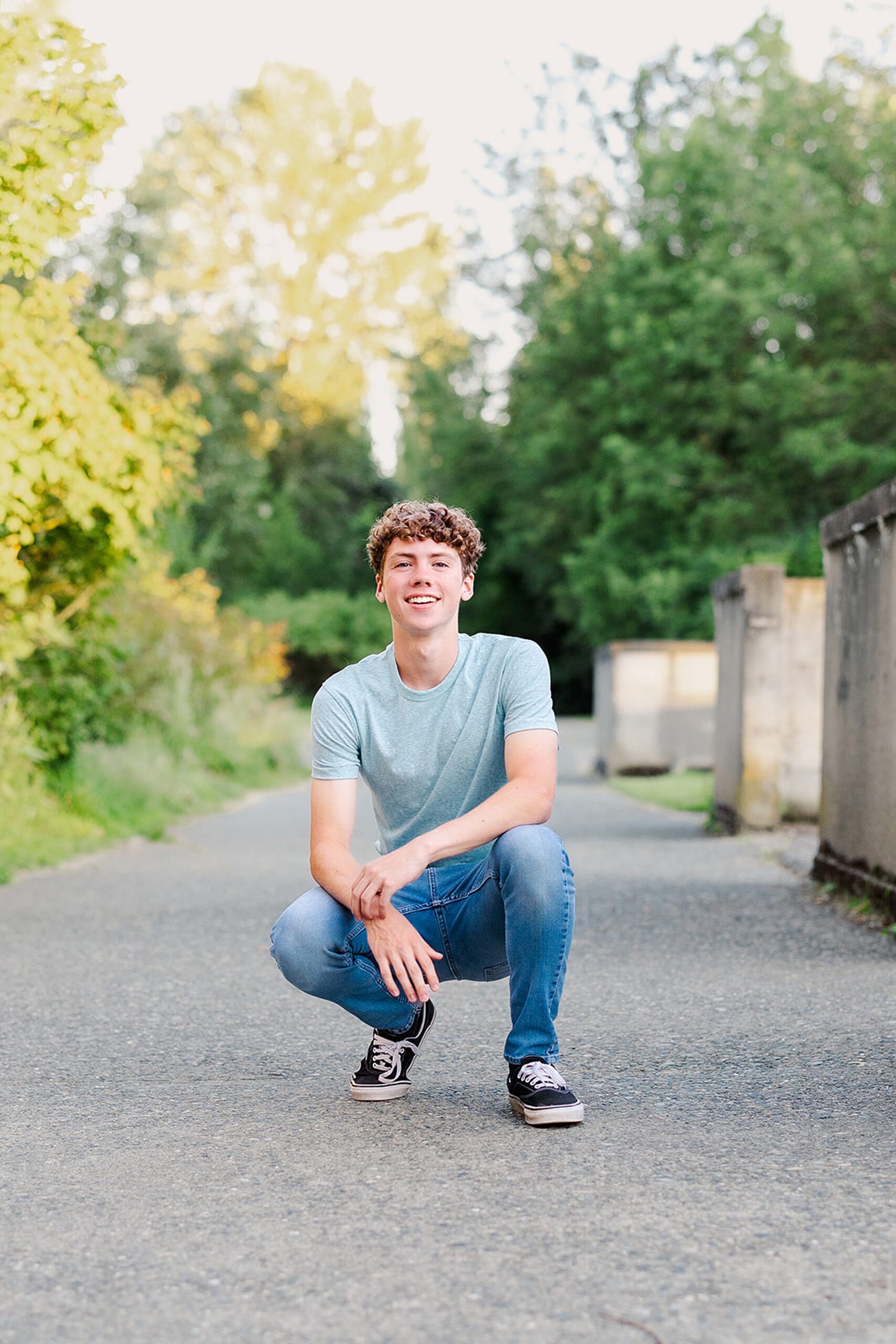 A teenage boy in jeans and a blue shirt squats in a park sidewalk at sunset before visiting colleges near bellevue wa