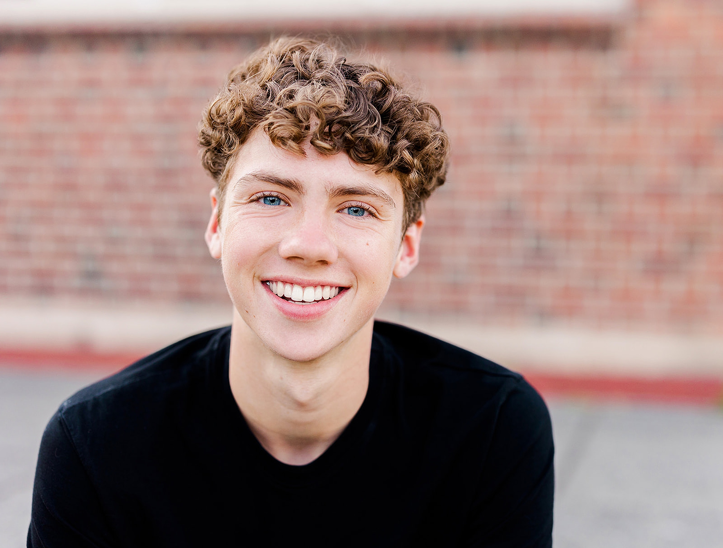 A smiling high school senior boy with curly hair sits by a brick wall in a black shirt before visiting colleges near bellevue wa